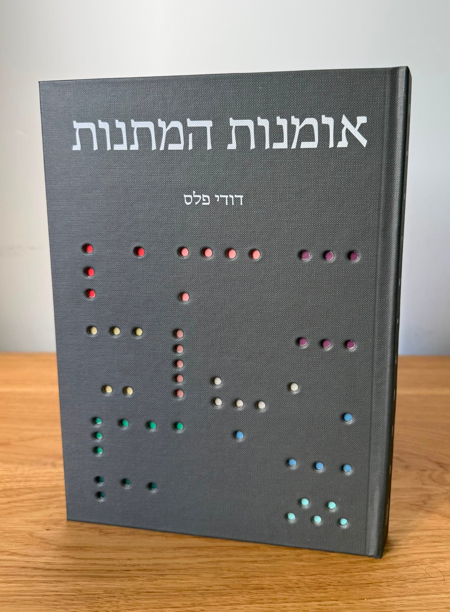 הספר ‘אומנות המתנות’ מאת דודי פלס עומד על שולחן עץ — כריכה קשה אפורה ועליה הכותרת ונקודות צבעוניות שקועות בבד